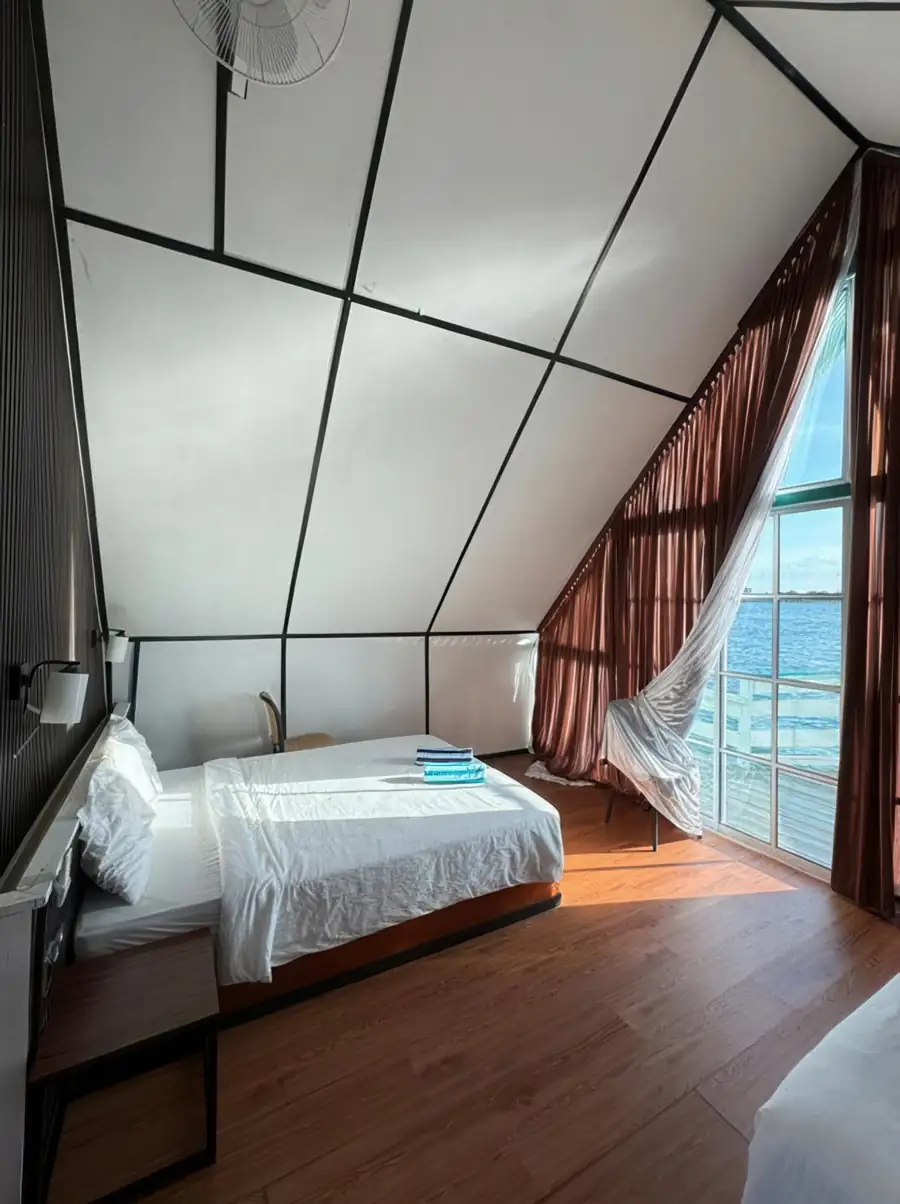 Sunlit bedroom interior at Adil Waterhouse Resort featuring a queen bed and an air conditioning unit with large windows that frame the marine sanctuaries of the Celebes Sea.